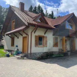 Czyszczenie elewacji domu dwukondygnacyjnego z elementami drewnianymi, widoczny pracownik na rusztowaniu i ślady mycia ciśnieniowego.