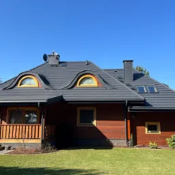 Dom z ciemnoszarym dachem z dachówki bitumicznej i drewnianą elewacją w ciepłym odcieniu brązu. Widoczne okna z żółtymi ramami i zadbany trawnik.