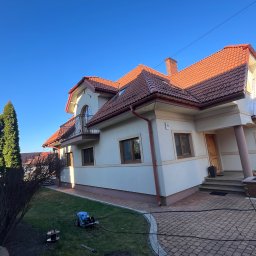 K-BUD Krystian Ujazdowski - Dwupiętrowy dom z czerwonym dachem i beżową elewacją w słoneczny dzień. Na trawniku sprzęt do mycia ciśnieniowego. Kostka brukowa wokół domu.