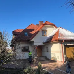 Pracownik w jaskrawej kamizelce myje dach domu z czerwonej dachówki myjką ciśnieniową. Część dachu jest już czysta, kontrastując z zabrudzoną resztą. Błękitne niebo w tle.