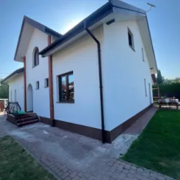 Biały dom z ciemnobrązowymi akcentami, widoczny z perspektywy ogrodu. Kostka brukowa prowadzi do wejścia. Słońce za budynkiem tworzy subtelną aurę.