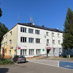 K-BUD Krystian Ujazdowski - Trzykondygnacyjny budynek z białą elewacją i czarnym dachem, antena na dachu, widoczny fragment parkingu i drzewa. Budynek z rampą dla niepełnosprawnych.