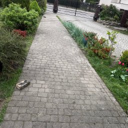 K-BUD Krystian Ujazdowski - Długa, szara ścieżka z kostki brukowej prowadzi do metalowej bramy. Po bokach ścieżki rosną krzewy i trawa. Na kostce leży pojedyncza cegła. W tle domy.