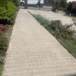 K-BUD Krystian Ujazdowski - Ułożona szara kostka brukowa tworząca ścieżkę w ogrodzie, prowadząca do bramy wjazdowej z częściowo widocznym samochodem dostawczym w tle.