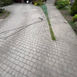 K-BUD Krystian Ujazdowski - Szara kostka brukowa na podjeździe, z widocznym wężem ciśnieniowym i pistoletem do mycia. Częściowo umyta powierzchnia. Otoczenie: trawnik i roślinność.