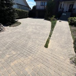 K-BUD Krystian Ujazdowski - Szeroki kadr na ułożoną kostkę brukową prowadzącą do domu z białymi ścianami. Po prawej stronie trawnik z krzewami, po lewej drzewo iglaste. W tle sąsiedni dom.