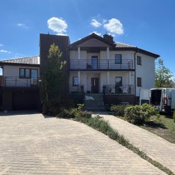 K-BUD Krystian Ujazdowski - Dwupiętrowy dom z balkonami, kostką brukową i psem na schodach wejściowych. Widoczny samochód dostawczy z otwartymi drzwiami. Słoneczny dzień, błękitne niebo.