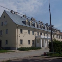 K-BUD Krystian Ujazdowski - Długi budynek wielorodzinny z szarym dachem w Zabłudowie. Kremowa elewacja, małe okna, widoczne anteny satelitarne. Ujęcie z poziomu ulicy.