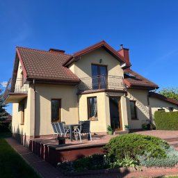 K-BUD Krystian Ujazdowski - Jasna elewacja domu z brązowym dachem i balkonami z ozdobnymi balustradami. Na tarasie zestaw mebli ogrodowych. Błękitne niebo w tle.