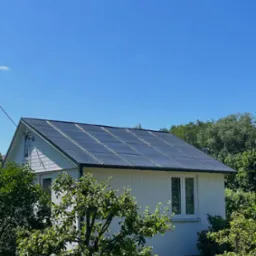 Biały dom z panelami słonecznymi na dachu, otoczony zielenią. Widok na fasadę i dach w słoneczny dzień. Panele równomiernie rozmieszczone, tworzą nowoczesny wygląd.