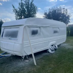 Przyczepa kempingowa po odświeżeniu koloru na biało, z zabezpieczonymi oknami i kołami, na trawniku z widocznymi śladami farby w tle drzewa i niebo.