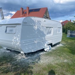 K-BUD Krystian Ujazdowski - Przyczepa kempingowa po świeżym malowaniu na biało, z zabezpieczonymi oknami folią, na tle zielonego trawnika i domu z czerwoną dachówką w pochmurny dzień.