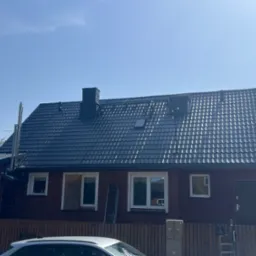 Dom z ciemną, nową dachówką i kominem. Przy ścianie drabina, okno zabite deskami. Widoczne elementy elewacji i ogrodzenia. Słoneczny dzień.