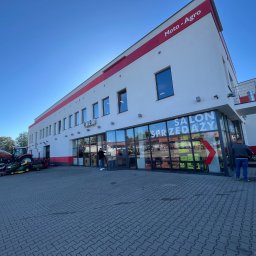K-BUD Krystian Ujazdowski - Biały budynek Moto-Agro z czerwonymi akcentami, salon sprzedaży z przeszkloną fasadą, kostka brukowa na zewnątrz i sprzęt rolniczy w tle. Błękitne niebo.