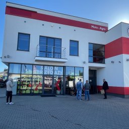 K-BUD Krystian Ujazdowski - Dwukondygnacyjny budynek z napisem 'Salon sprzedaży' w oknach, pomalowany na biało z czerwonymi akcentami. Przed budynkiem stoi kilka osób i zaparkowany samochód.