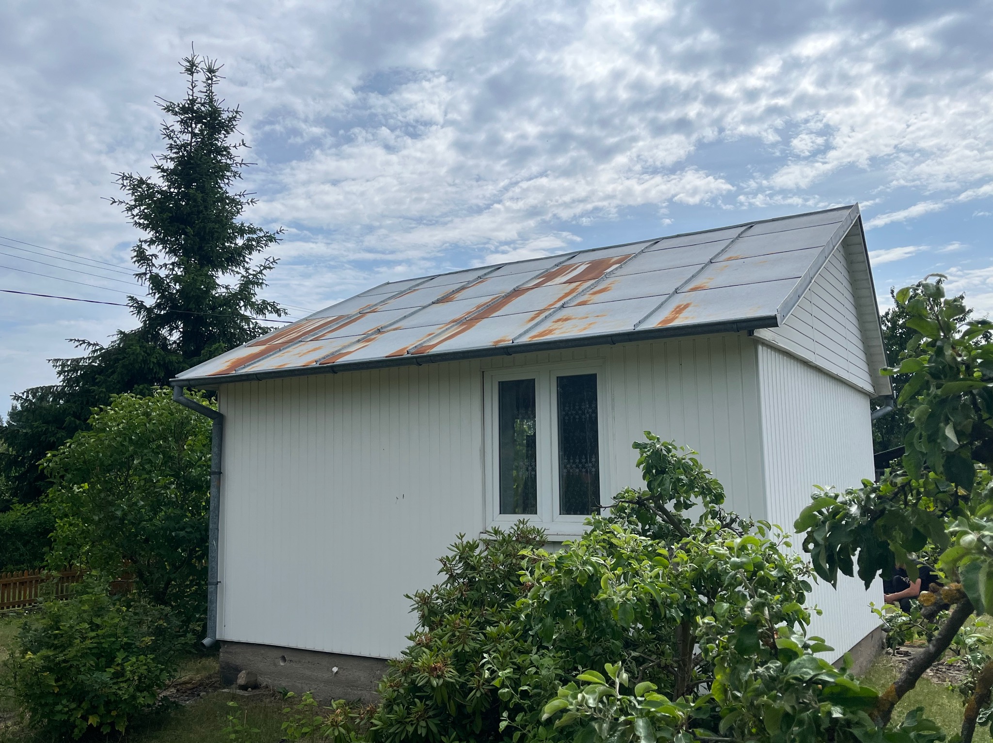 Biały budynek z blachy trapezowej z rdzawymi śladami na dachu, otoczony zielenią. Widok z dołu, pochmurne niebo w tle.