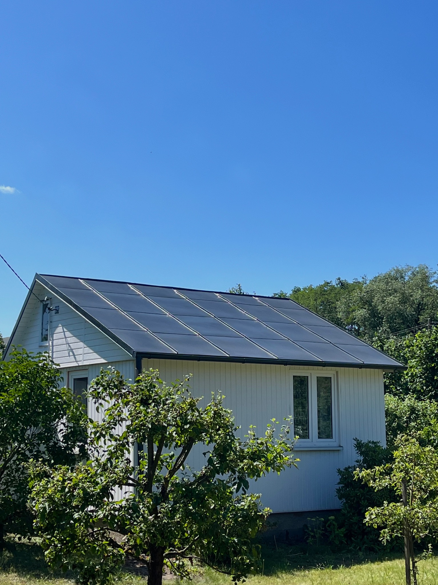 Biały dom z panelami słonecznymi na dachu, otoczony zielenią. Widok na fasadę i dach w słoneczny dzień. Panele równomiernie rozmieszczone, tworzą nowoczesny wygląd.