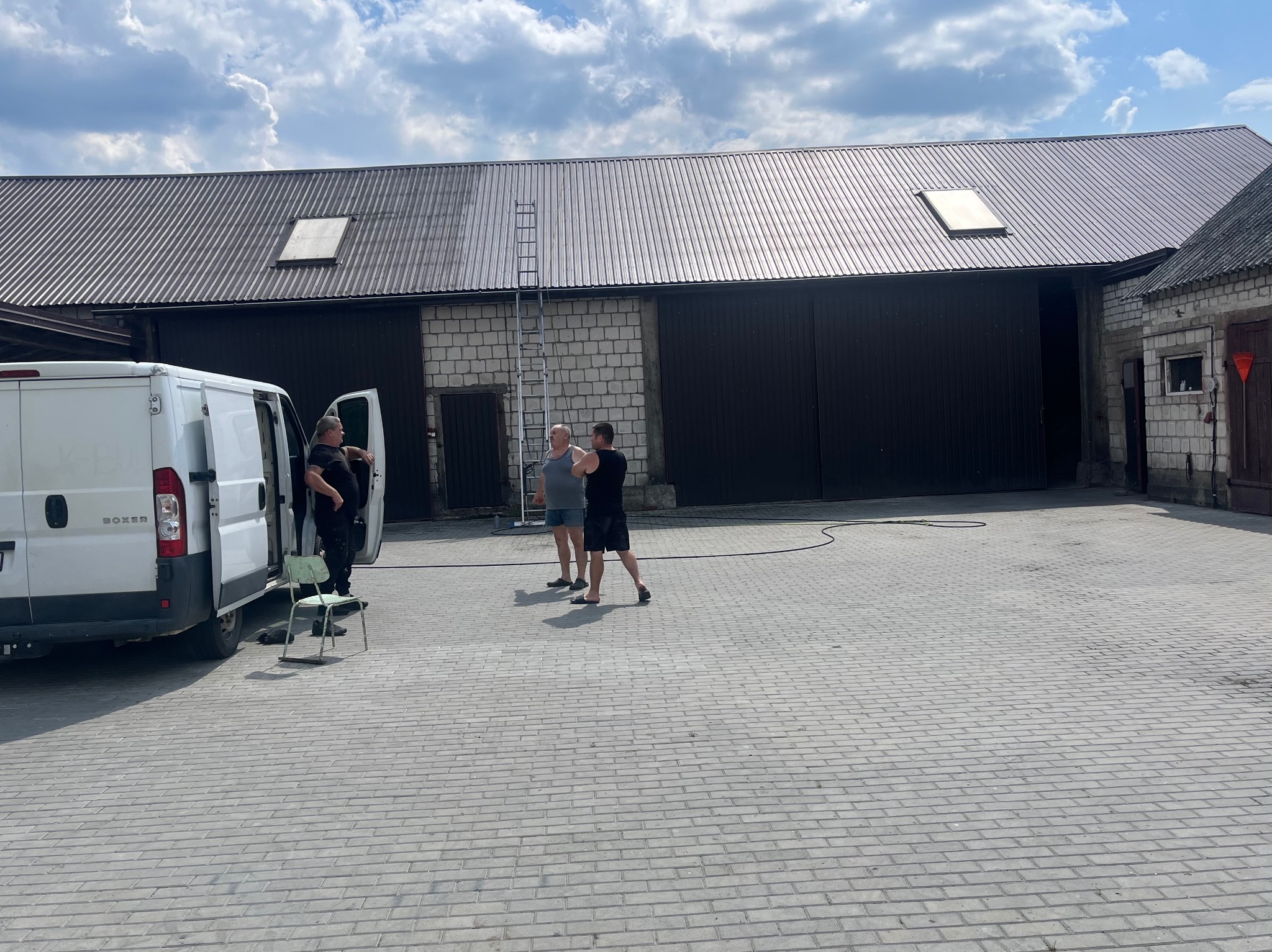 Częściowo umyty dach budynku gospodarczego z blachy trapezowej, widoczna drabina oparta o ścianę. Na pierwszym planie ekipa przy białym busie.