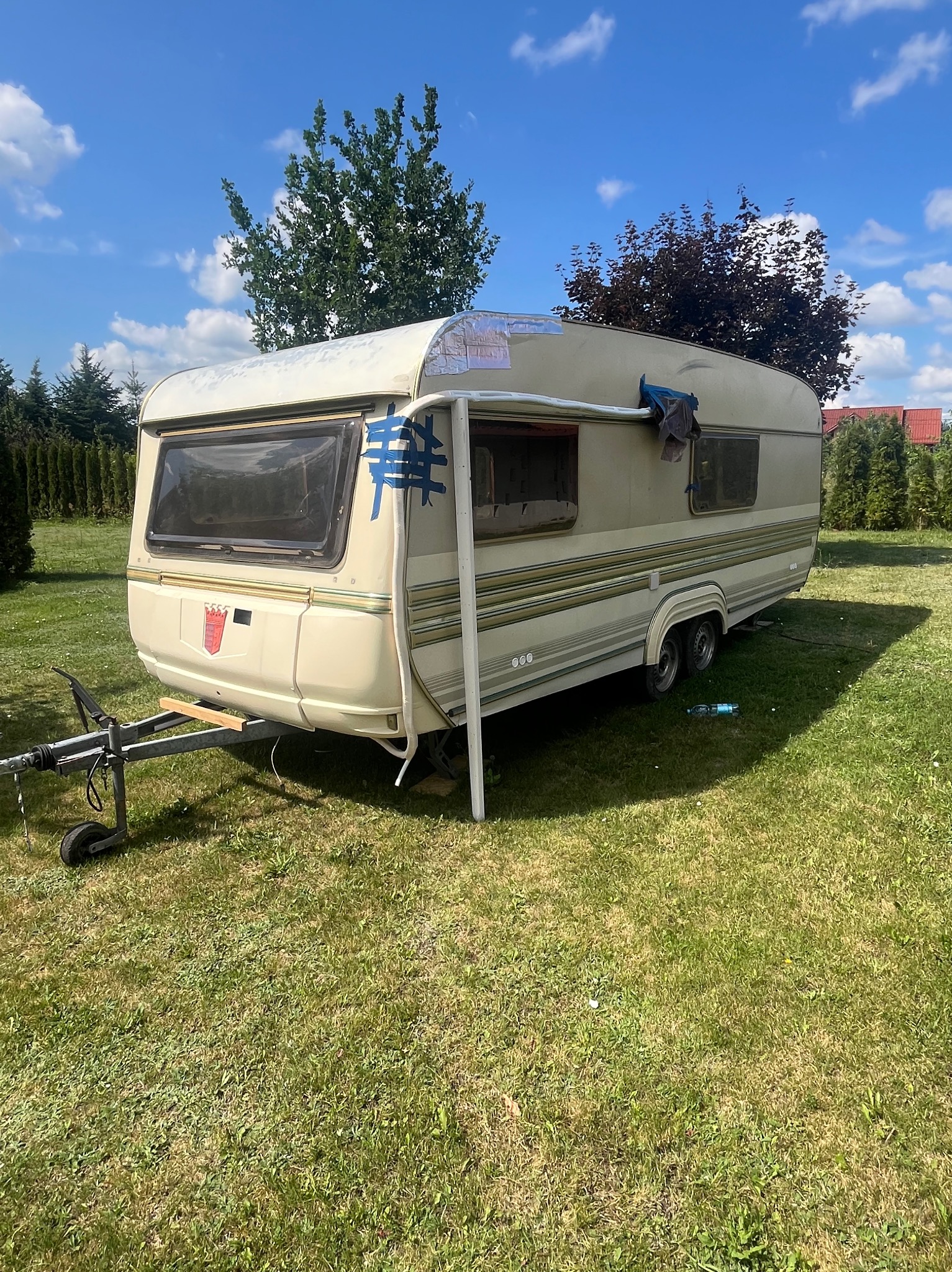 Kempingowa przyczepa na trawie, z widocznymi śladami użytkowania i niebieską taśmą. Tło stanowią drzewa i błękitne niebo z chmurami.