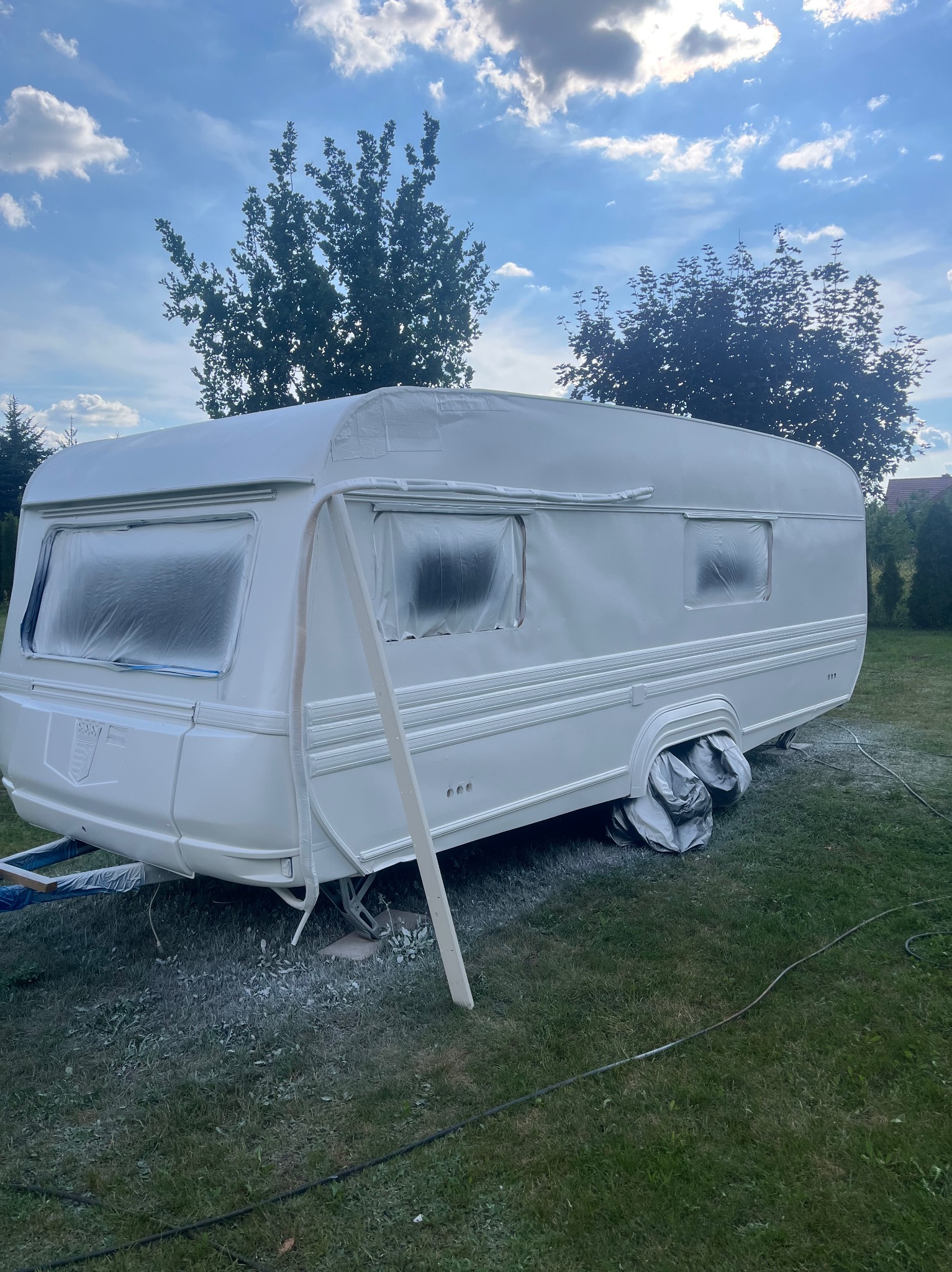 Przyczepa kempingowa po odświeżeniu koloru na biało, z zabezpieczonymi oknami i kołami, na trawniku z widocznymi śladami farby w tle drzewa i niebo.