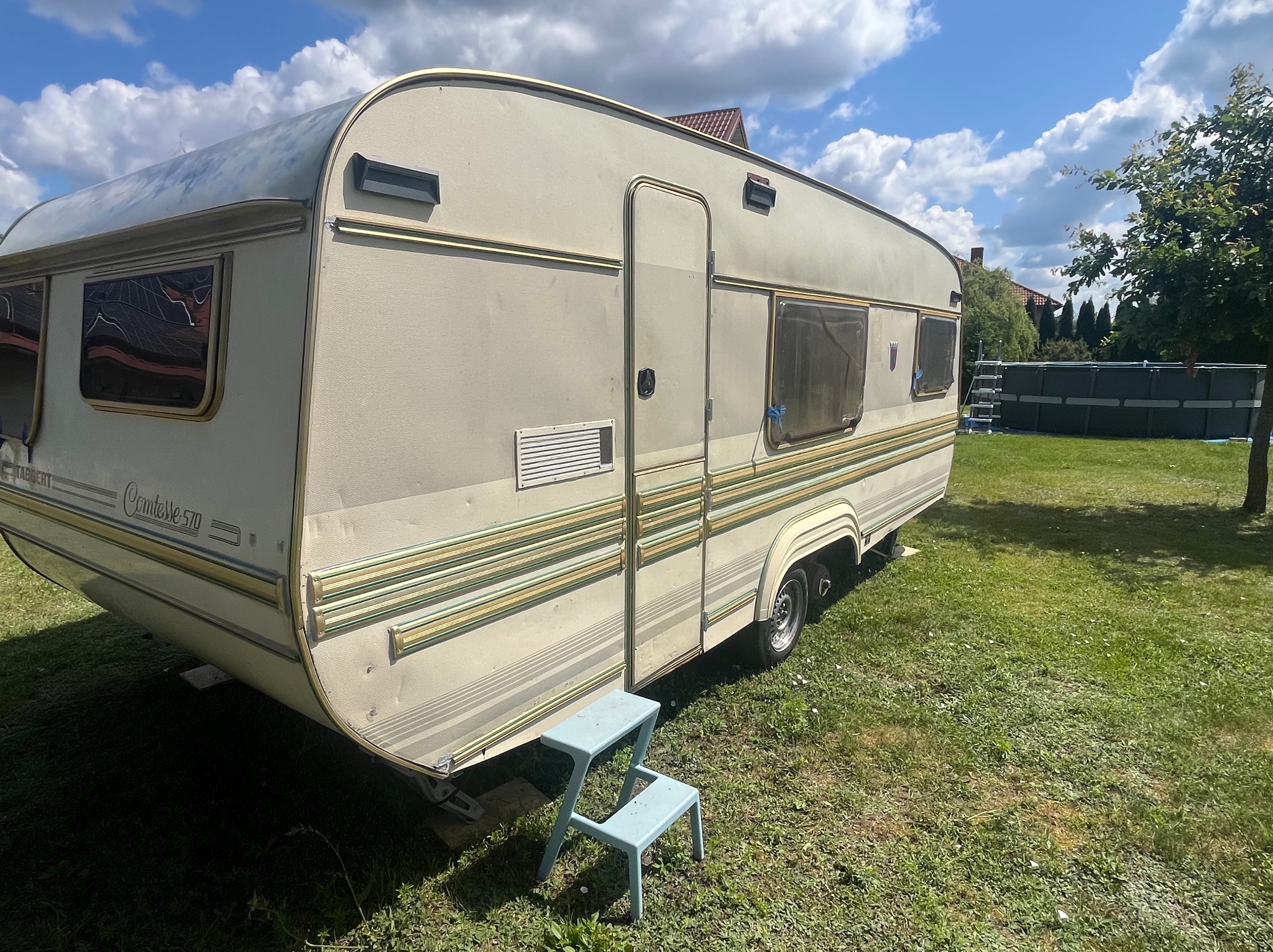 Przyczepa kempingowa Tabbert Comtesse 570 na trawie, z niebieskim stołkiem obok wejścia. W tle drzewa i basen ogrodowy.
