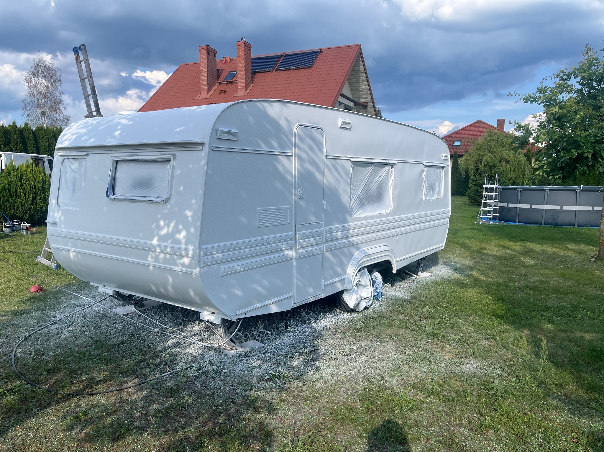 Przyczepa kempingowa po świeżym malowaniu na biało, z zabezpieczonymi oknami folią, na tle zielonego trawnika i domu z czerwoną dachówką w pochmurny dzień.