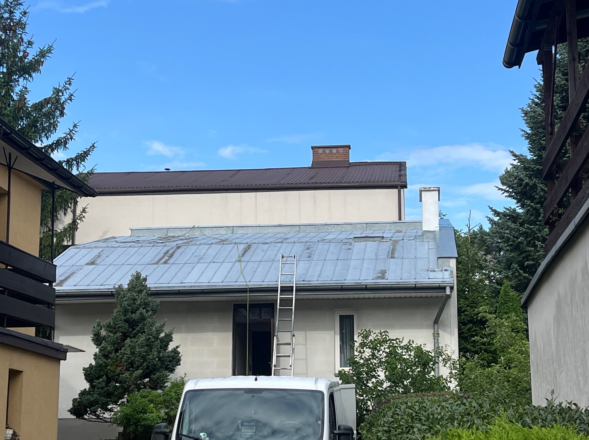 Dom z blachy trapezowej, drabina oparta o dach, widoczny fragment samochodu. Przygotowania do prac na wysokości, błękitne niebo w tle.