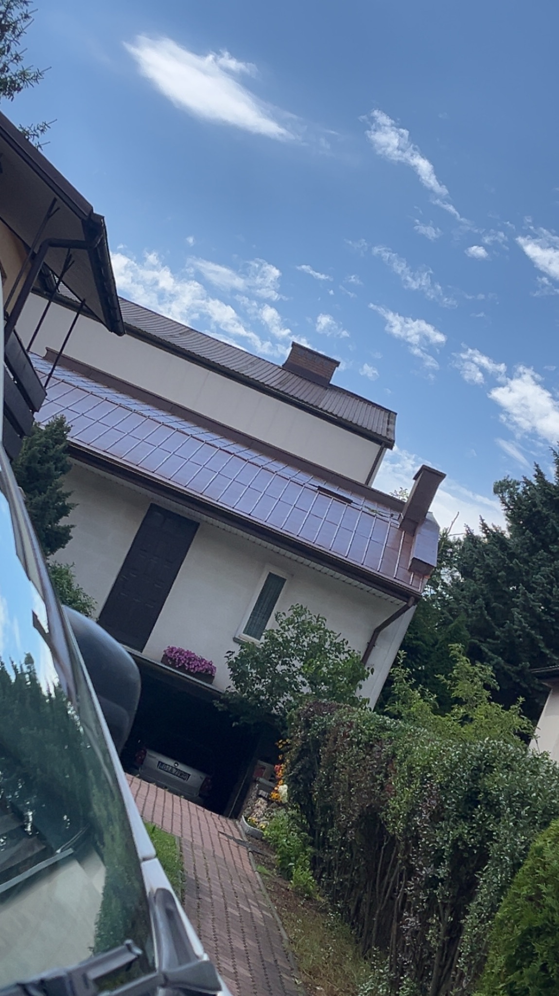 Dwupiętrowy dom z brązowym dachem i białymi ścianami, widziany z perspektywy ulicy, z fragmentem samochodu w kadrze. W tle błękitne niebo z chmurami.