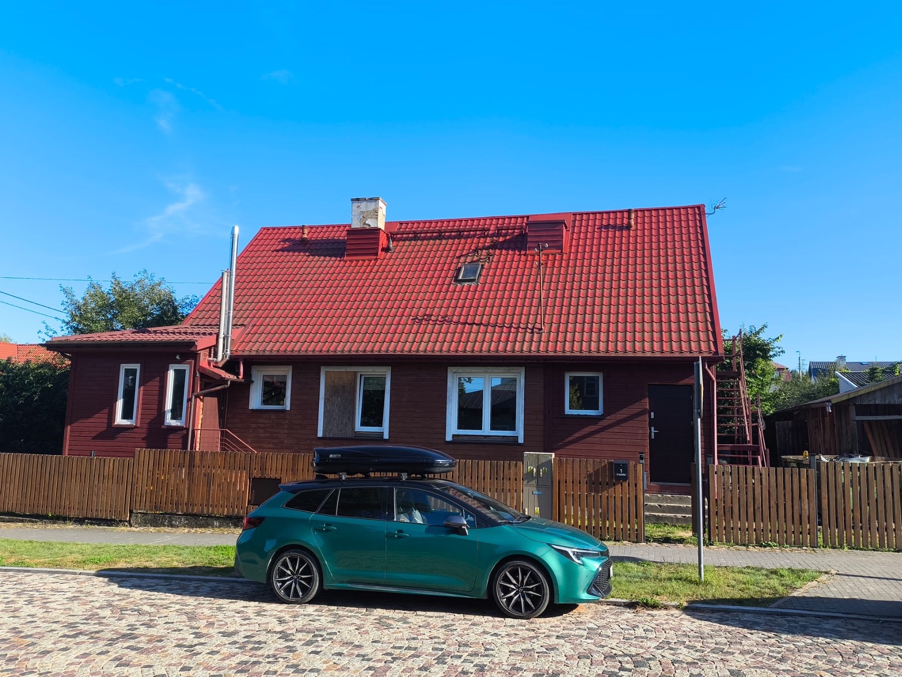 Ciemnobrązowy dom z czerwoną dachówką, widziany z poziomu kostki brukowej. Na pierwszym planie zielony samochód z bagażnikiem dachowym.