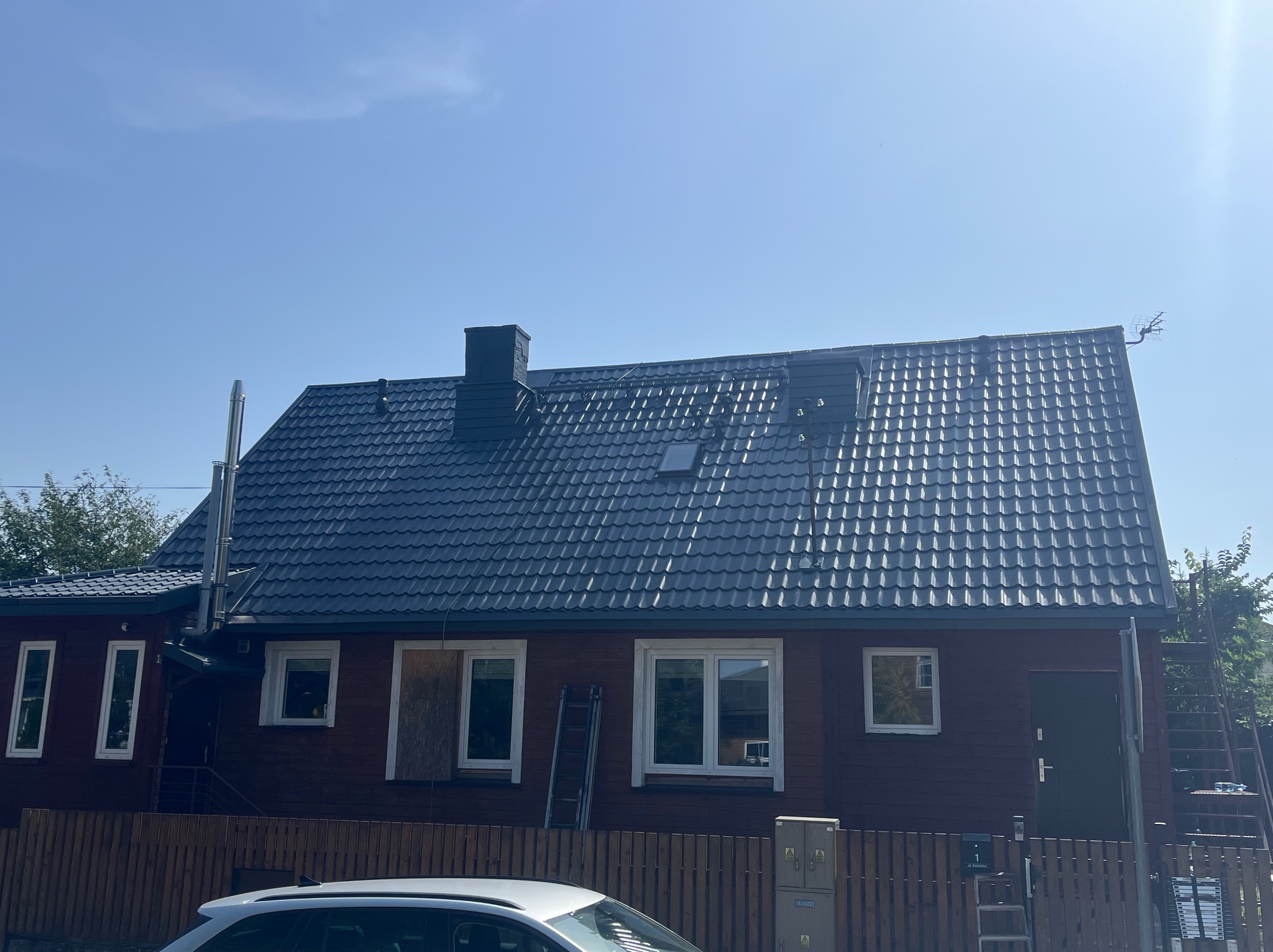 Dom z ciemną, nową dachówką i kominem. Przy ścianie drabina, okno zabite deskami. Widoczne elementy elewacji i ogrodzenia. Słoneczny dzień.