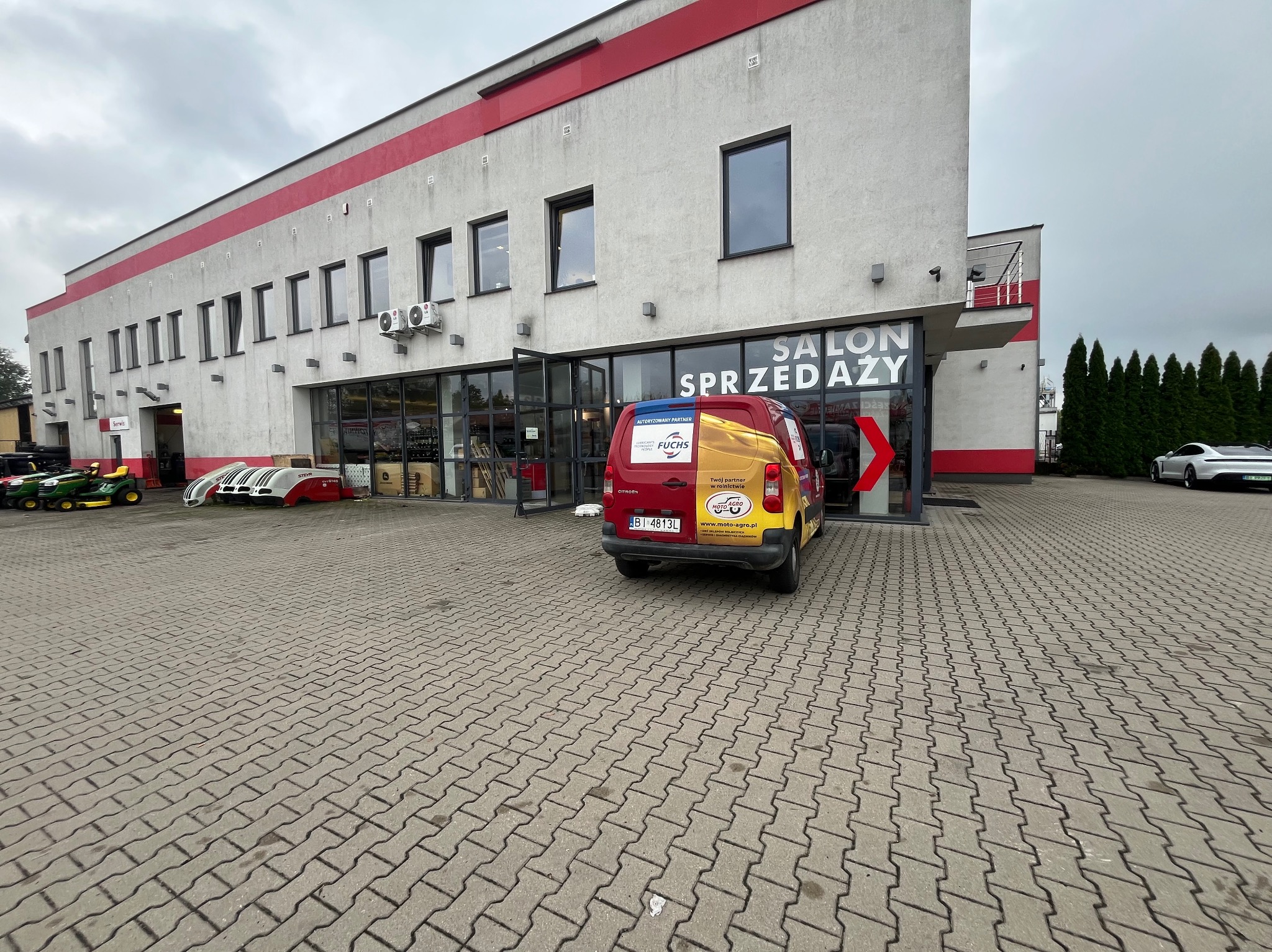 Budynek salonu sprzedaży z czerwonymi akcentami, zaparkowany firmowy van z logo Moto-Agro i Fuchs, kostka brukowa na pierwszym planie, pochmurne niebo.