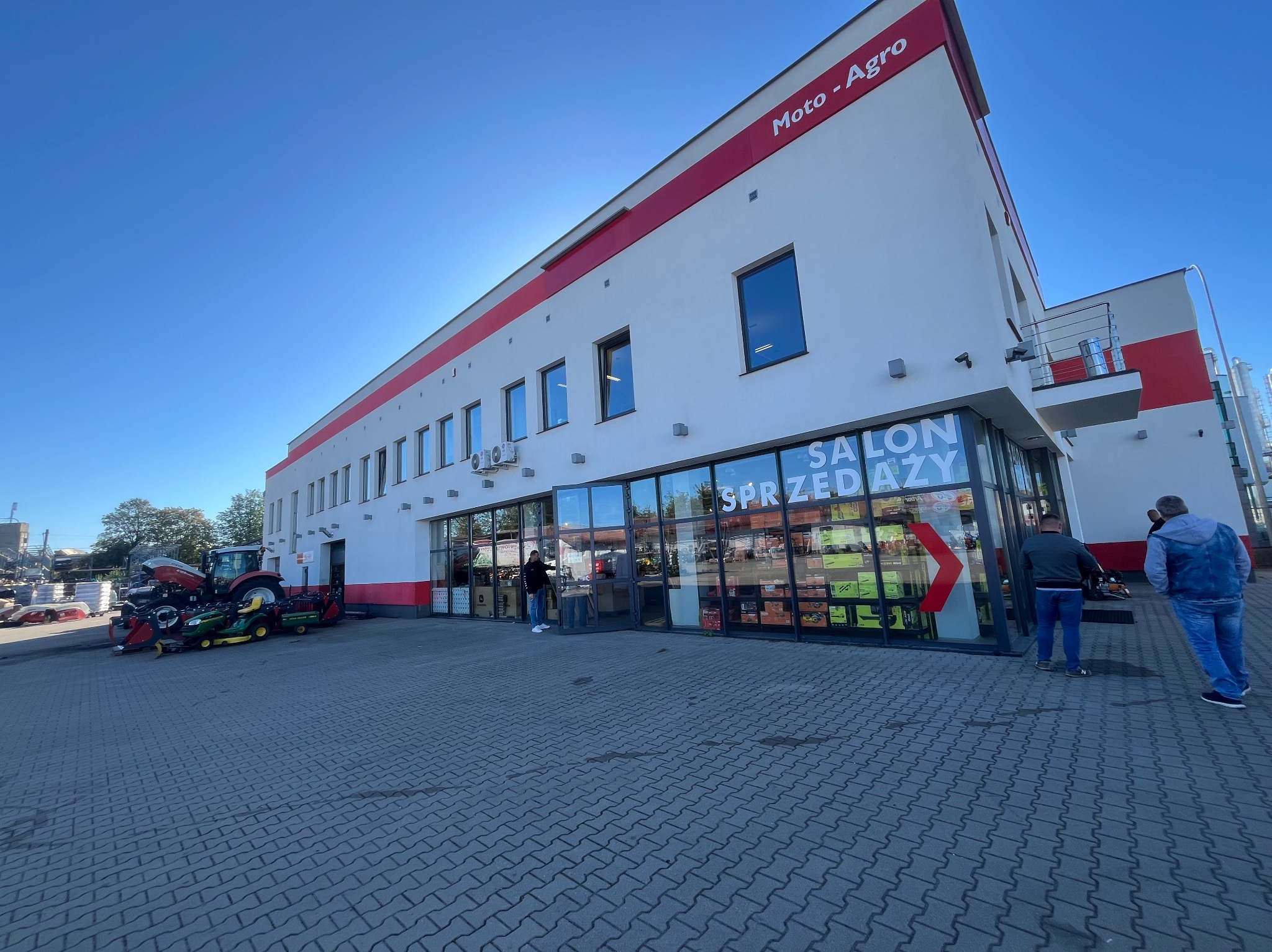 Biały budynek Moto-Agro z czerwonymi akcentami, salon sprzedaży z przeszkloną fasadą, kostka brukowa na zewnątrz i sprzęt rolniczy w tle. Błękitne niebo.