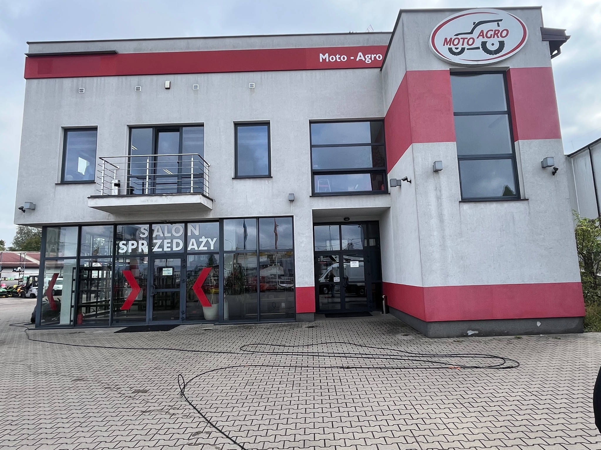 Dwukondygnacyjny budynek Moto-Agro w Zabłudowie z balkonem i dużymi oknami, widoczny z poziomu kostki brukowej. Na elewacji logo firmy i napis Salon Sprzedaży.