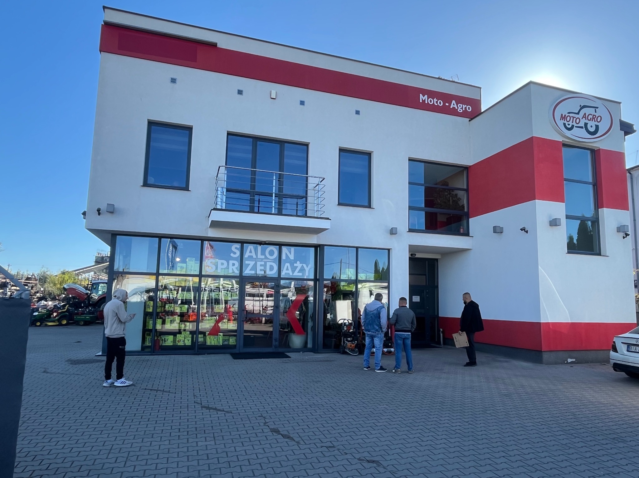 Dwukondygnacyjny budynek z napisem 'Salon sprzedaży' w oknach, pomalowany na biało z czerwonymi akcentami. Przed budynkiem stoi kilka osób i zaparkowany samochód.
