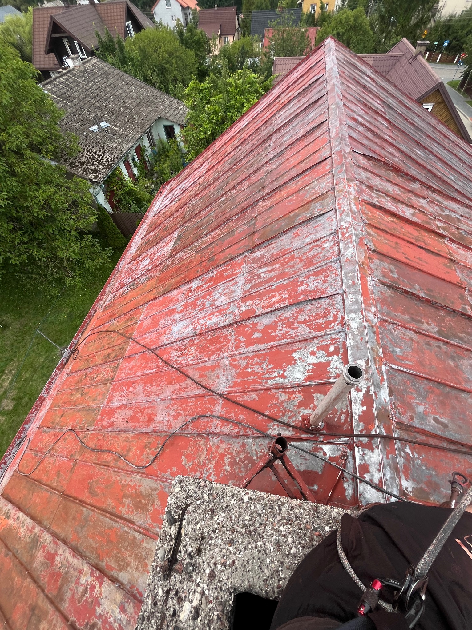 Czerwony, metalowy dach z widocznymi śladami korozji i ubytkami farby, sfotografowany z góry. Widoczny komin i fragment uprzęży zabezpieczającej.