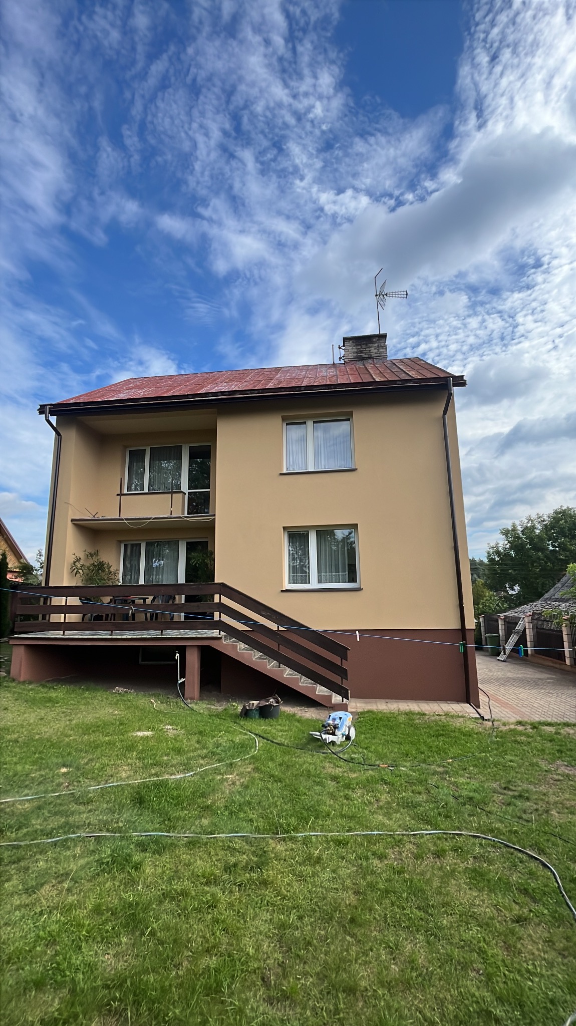 Dwupiętrowy dom z beżową elewacją i czerwoną dachówką, widoczny z poziomu trawnika. Na trawie leży wąż i myjka ciśnieniowa. Błękitne niebo z chmurami w tle.