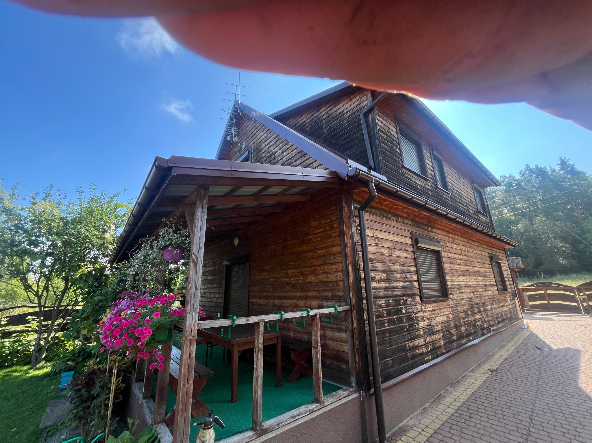 Drewniany dom z werandą, widok z dołu, błękitne niebo. Na werandzie kwiaty w donicach, zielona podłoga. Widoczna elewacja i fragment kostki brukowej.