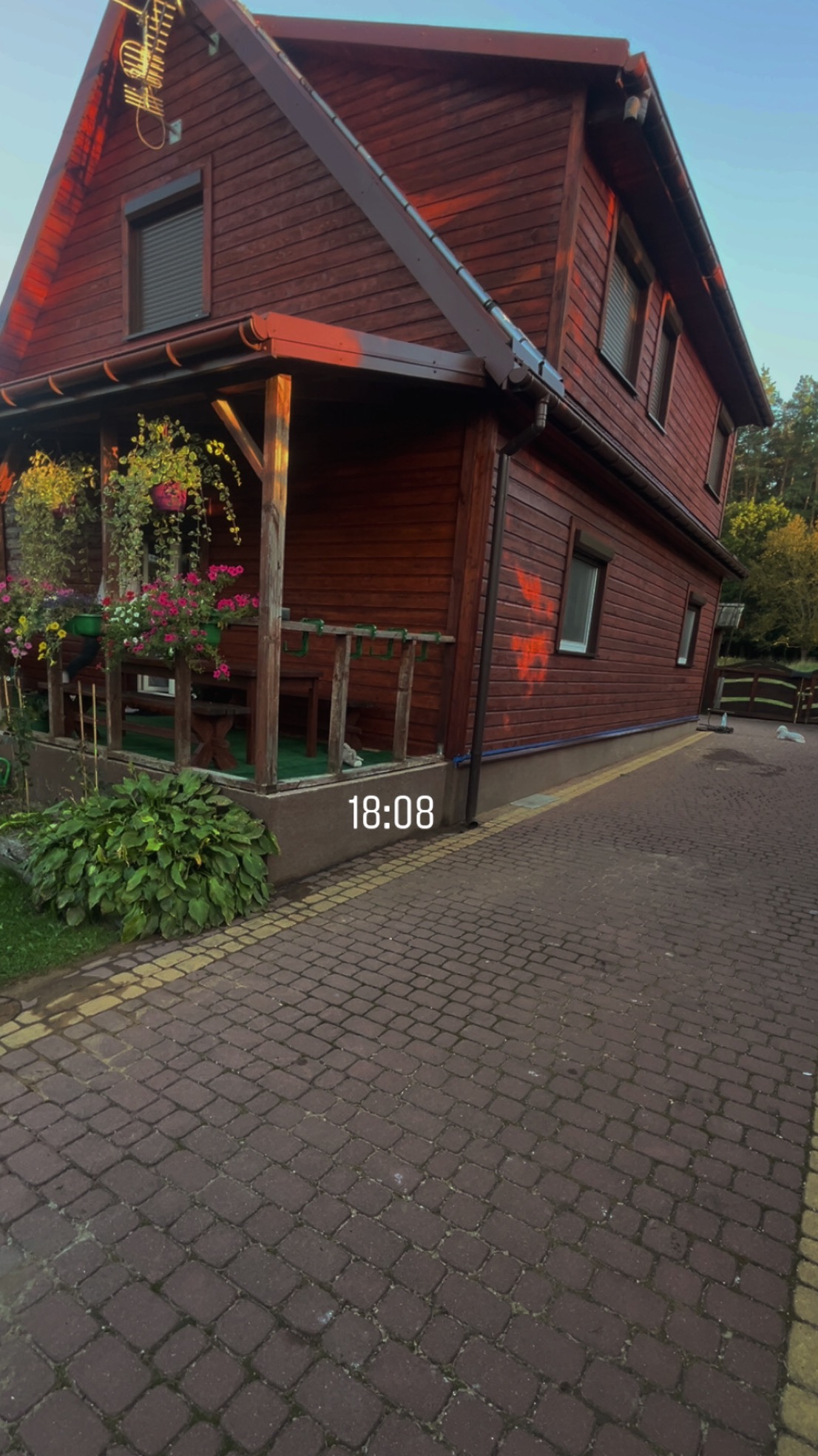 Drewniany dom z werandą, ozdobiony kwiatami w donicach. Czerwona kostka brukowa prowadzi do wejścia. Widoczne okna z roletami i fragment dachu. Ciepłe, wieczorne światło.
