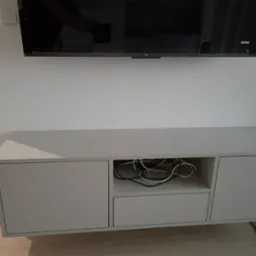 Biała, wisząca szafka RTV z dwiema szafkami zamykanymi i otwartą półką na kable, zamontowana pod telewizorem na białej ścianie.