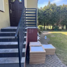 Nowoczesna balustrada schodowa z czarnych, prostych profili, widok na wejście do domu i stosy płytek na posesji.