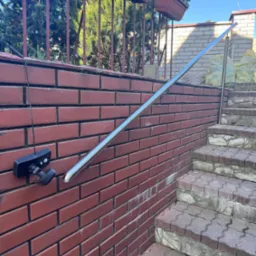 Nierdzewna poręcz zamontowana na ceglanym murze przy schodach zewnętrznych. Na murze widoczna lampa. W tle roślinność i worki z materiałami budowlanymi.