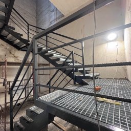 professional mobile welder PMWelders - Metalowe schody z platformą kratową w surowym wnętrzu. Widoczne spawy i elementy konstrukcyjne, oświetlenie lampą. Kąt ujęcia od dołu.
