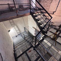 professional mobile welder PMWelders - Metalowe schody kratowe z czarną poręczą w surowym wnętrzu, widok z góry. Na stopniach widoczne narzędzia i osprzęt. Fragment ceglanej ściany w tle.