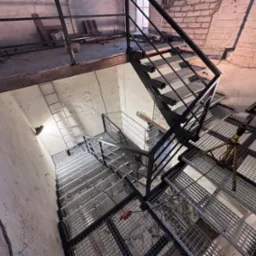 Metalowe schody kratowe z czarną poręczą w surowym wnętrzu, widok z góry. Na stopniach widoczne narzędzia i osprzęt. Fragment ceglanej ściany w tle.