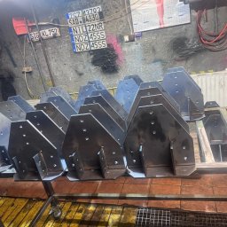 professional mobile welder PMWelders - Metalowe elementy konstrukcyjne, prawdopodobnie wsporniki, ułożone na wózku warsztatowym w surowym wnętrzu. Widoczne ślady obróbki i otwory montażowe.