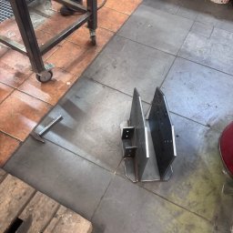 professional mobile welder PMWelders - Metalowa konstrukcja spawana z dwóch pionowych płyt, leżąca na posadzce warsztatu, obok młotek i fragmenty stołu spawalniczego na kółkach.