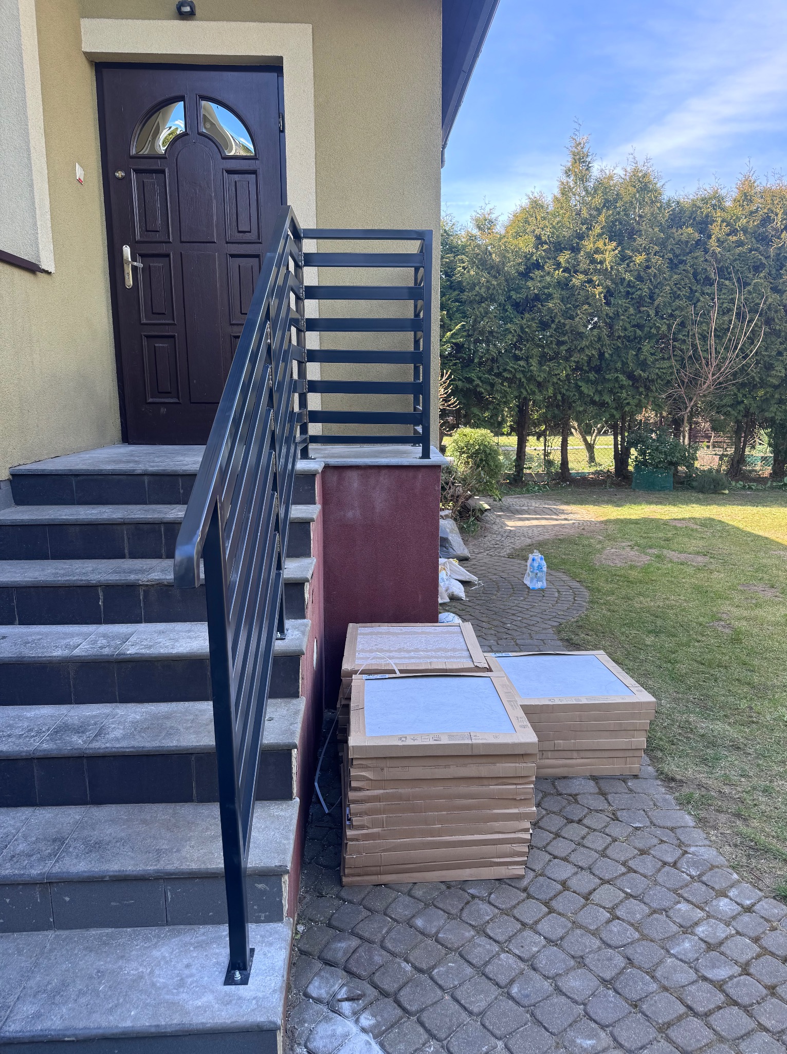 Nowoczesna balustrada schodowa z czarnych, prostych profili, widok na wejście do domu i stosy płytek na posesji.