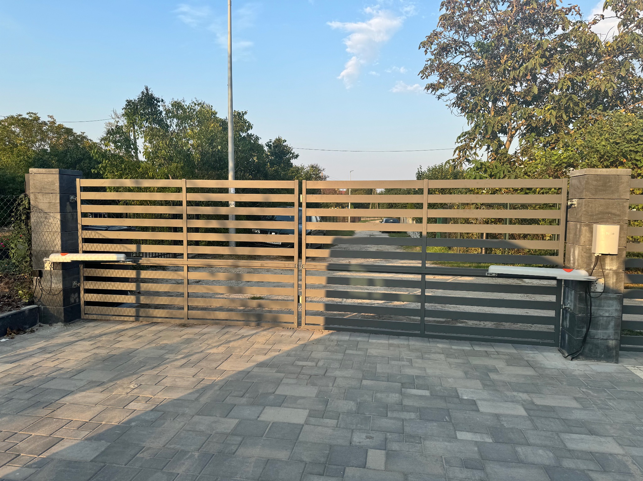 Automatyczna brama wjazdowa z poziomymi przęsłami, zamontowana na betonowych słupkach, z widocznym napędem i kostką brukową na podjeździe.