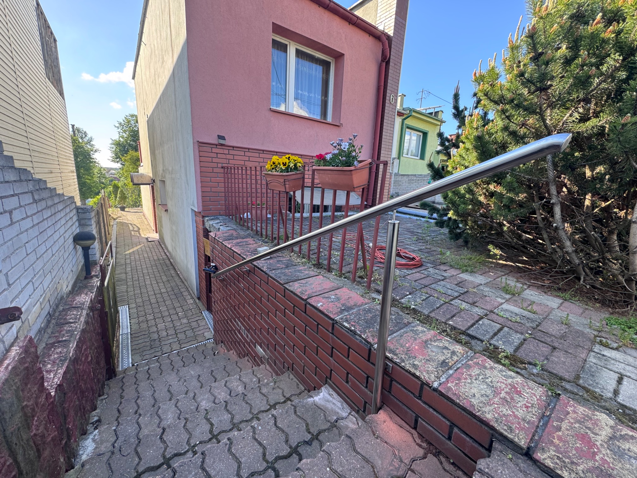 Zewnętrzne schody z szarej kostki brukowej, zabezpieczone niską, czerwoną ceglaną ścianką i metalową poręczą, prowadzące w dół wzdłuż budynku.