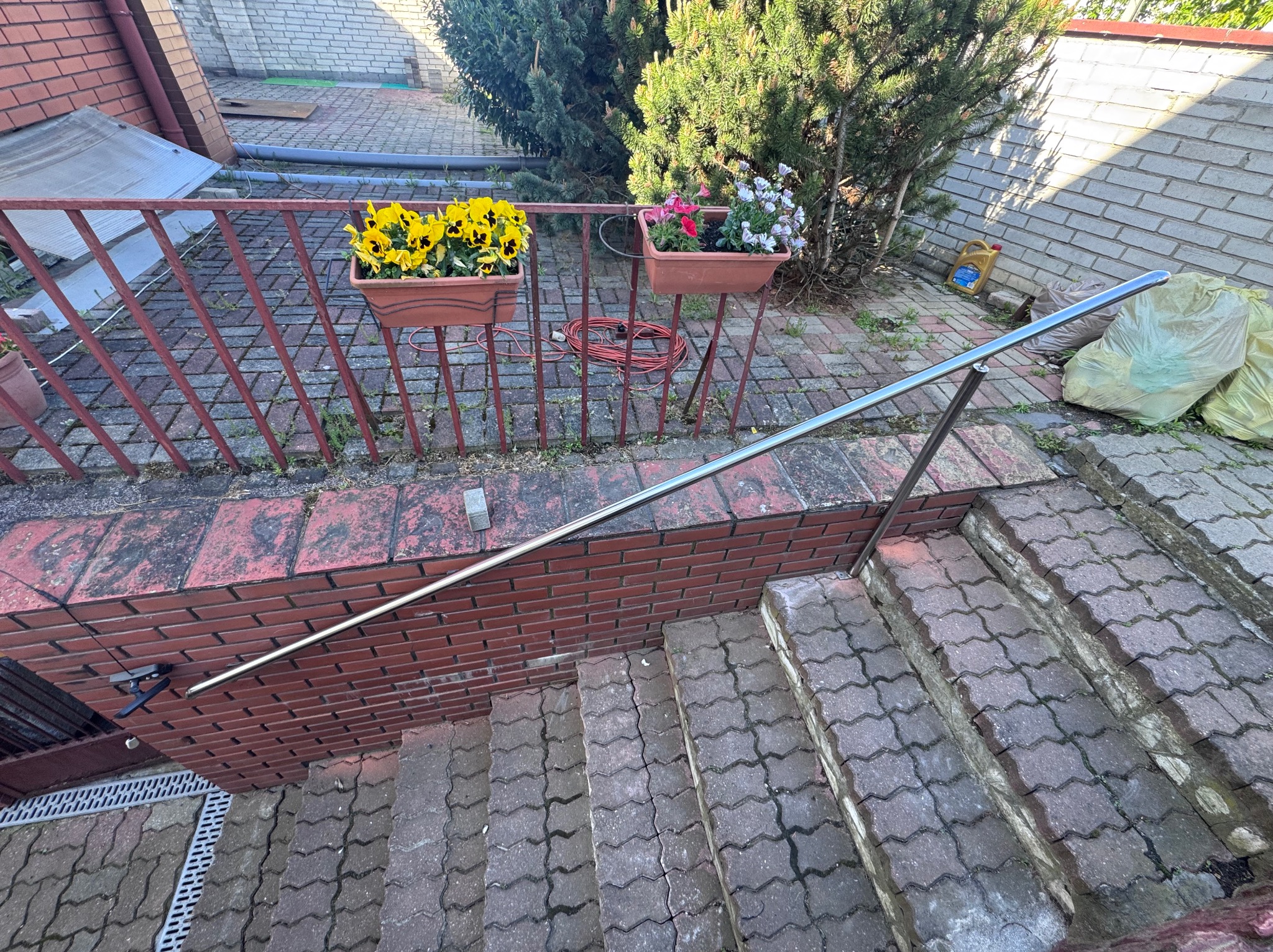 Zewnętrzne schody z poręczą ze stali nierdzewnej, prowadzące w dół. Czerwona, metalowa barierka z kwiatami w donicach. Kostka brukowa na schodach i podłożu.